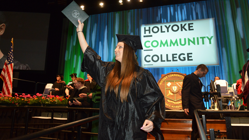 A smiling female HCC graduate wearing her cap and gown, holding her diploma at Commencement