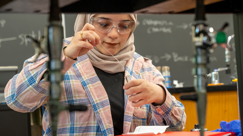 A student wearing protective eyewear works with a flame over a bunson burner