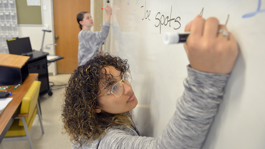 Female student wearing glasses and writing on a whiteboard in class