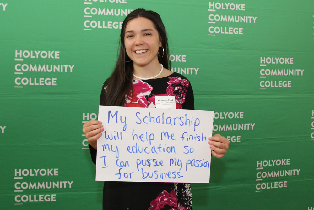 A student holds a handwritten sign reading "My scholarship will help me finish my education so I can pursue my passion for business."