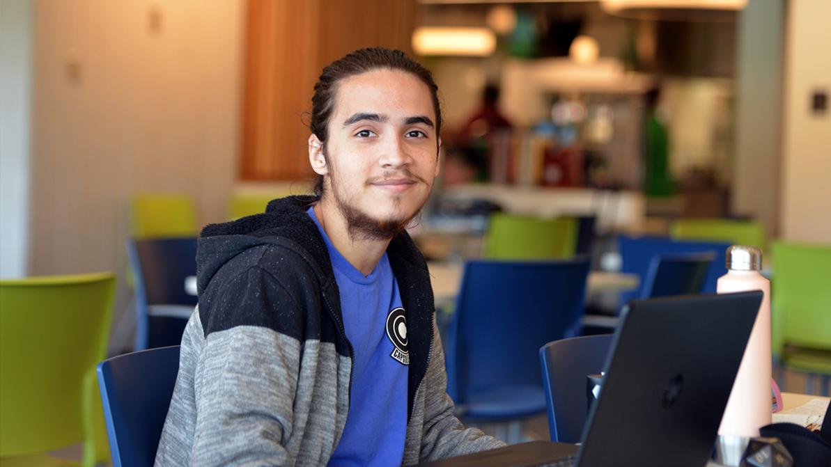 A student sits at a laptop computer. They smile at the camera. They are wearing a gray jacket and blue t-shirt, and have dark facial hair.